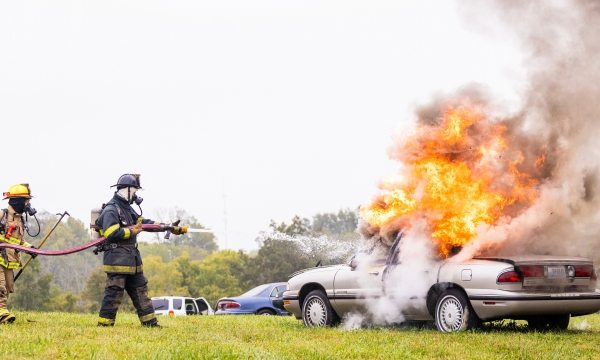 Two fire fighters tend to flames on a gray car, using a firehose.