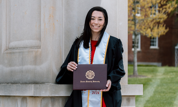 EKU graduate, Emma Thomas, holds her degree while wearing her graduation regalia
