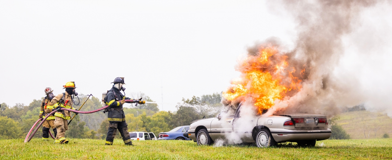 Two fire fighters tend to flames on a gray car, using a firehose.